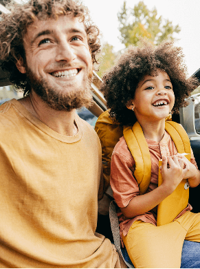 A smiling man and child sit in a car with the door open, the child wearing a yellow backpack.
