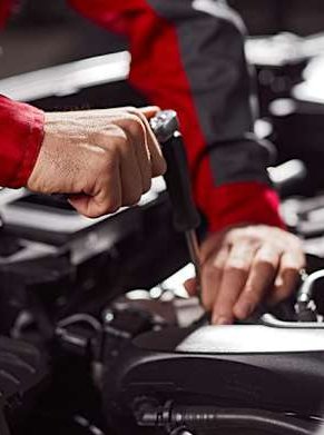 Mechanic in red and grey uniform working on a car engine with a tool.