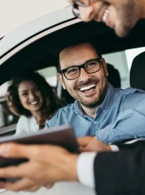Man and woman in car smiling at a person showing them a tablet.